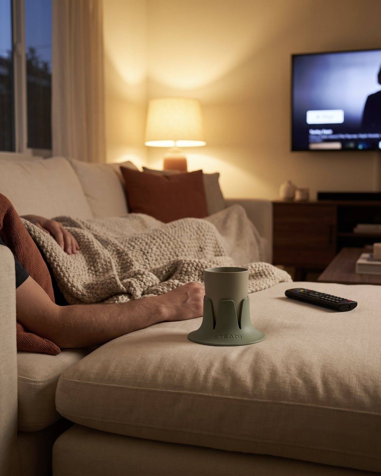 A sage drink holder cradling a tumbler on a couch cushion at dusk, TV glow behind.